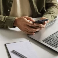 Uma pessoa utilizando um smartphone enquanto está em frente a um laptop em um ambiente de trabalho, com um bloco de anotações e caneta ao lado.