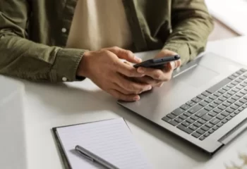 Uma pessoa utilizando um smartphone enquanto está em frente a um laptop em um ambiente de trabalho, com um bloco de anotações e caneta ao lado.