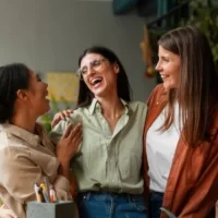 Três mulheres sorrindo e se divertindo em um ambiente leve e descontraído, cercadas por plantas e decoração moderna.